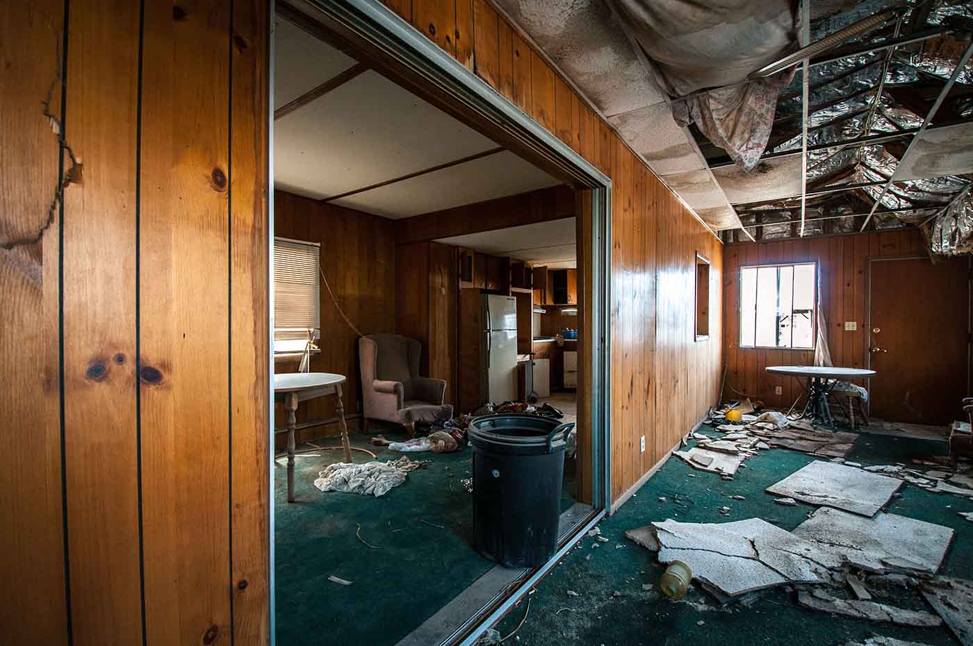 Interior of an abandoned house with wood-paneled walls, broken ceiling, and debris on the floor.